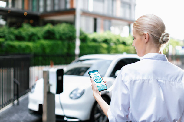 Suit-clad progressive businesswoman look at EV cars battery status from her phone while standing on a charging station with a power cable plug and a renewable energy-powered electric vehicle. - 写真・画像