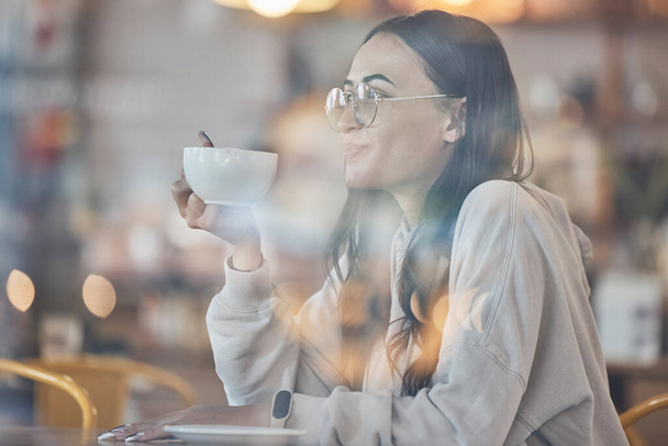 Thinking, smile and a woman drinking coffee in a cafe to relax alone over the weekend behind glass. Idea, happy and caffeine with an attractive young female enjoying a beverage in a restaurant. - Photo, image
