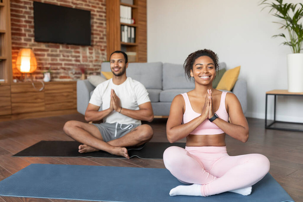 Glad young black couple in sportswear doing exercises, practicing yoga on mat, enjoy comfort, calm and silence, rest and relaxing in living room interior, copy space. Meditation at home, body care - Photo, Image
