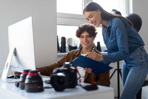 Женщина-ассистент с контрольным списком и мужчина-фотограф, работающий вместе на рабочем месте с компьютером и оборудованием, планируя фотосессию в студии - Фото, изображение