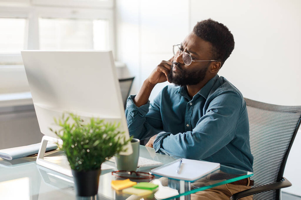 Gerente masculino negro pensativo sentado frente a la computadora, teniendo problemas, sentado en el escritorio en la oficina moderna y pensando. Concepto de temas de emprendimiento - Foto, imagen