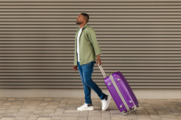 Usmívající se Afroameričan Muž Cestovatel Walking With Travel Suitcase Looking Aside Near Airport Wall Outdoors. Chlápek, co cestuje na dovolenou. Nabídka prohlídky a dopravy. Plná délka - Fotografie, Obrázek