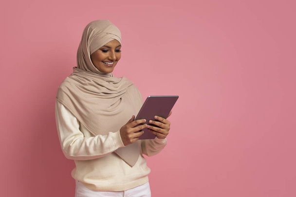 Usmívající se muslimská žena v hidžábu pomocí digitální tablety, nakupování on-line nebo prohlížení sociálních sítí, arabské ženy v šátku se těší moderní technologie, stojící nad růžové studio na pozadí - Fotografie, Obrázek