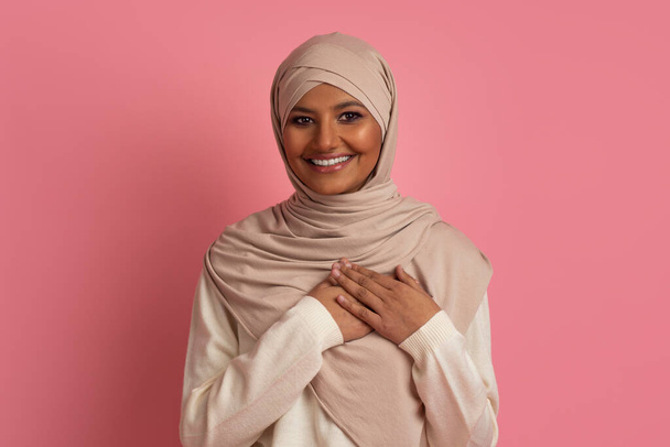 Concepto de gratitud. Retrato de mujer musulmana agradecida en hijab manteniendo las manos en el pecho y mirando a la cámara, sonriente dama árabe expresando aprecio, de pie sobre fondo de estudio rosa - Foto, Imagen