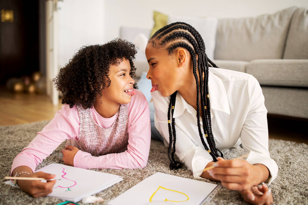 Grappige Afro-Amerikaanse mama en haar tienerdochter hebben plezier uitsteken tongen tekenen samen liggend op de vloer thuis. Familie band op het weekend. Hobby en vrijetijdsconcept - Foto, afbeelding