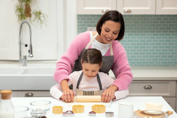 Onnellinen valkoihoinen nuori äiti auttaa pientä tytärtä esiliinoissa tekemään taikinaa evästeille, joissa on valssaustappi keittiön sisustuksessa. Tuki, kokki ruokaa, kotitekoisia makeisia, suhteita kotona - Valokuva, kuva