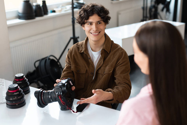 Cheerful male photographer talking with lady model, discussing result of photoshoot, sitting at desk with camera and lens in modern photostudio - Фото, изображение