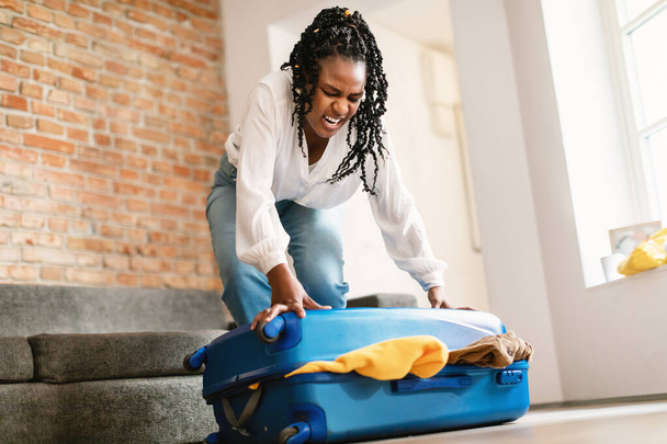 Glückliche afrikanisch-amerikanische Frau bereitet sich auf die Urlaubsreise vor, packt die Sachen und versucht, voller Kleider Koffer zu Hause im Wohnzimmer Innenraum zu schließen, freier Raum - Foto, Bild