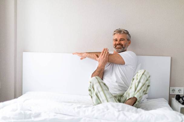 Joyful positive happy well-rested attractive grey-haired bearded mature man wearing pajamas sitting in bed at home, stretching arms and smiling, exercising after waking up, looking at copy space - Photo, Image