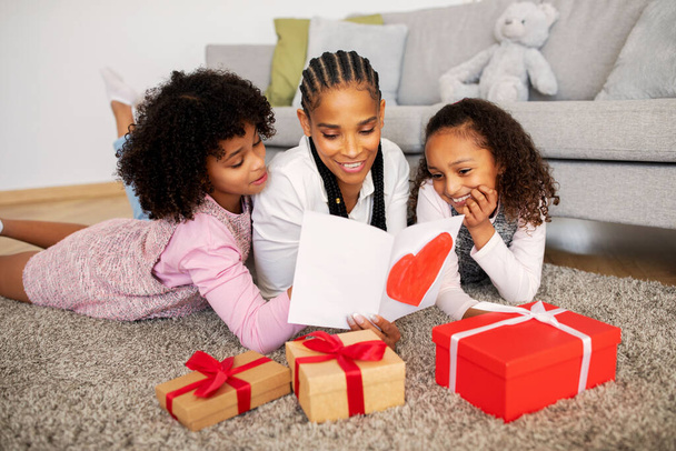 Nuori musta äiti ja kaksi tytärtä lukeminen onnittelukortti äitienpäivänä, lapset onnittelevat äitiään juhlimaan perhelomaa tai syntymäpäiväänsä lattialla käärittyjen lahjapakkausten joukossa kotona - Valokuva, kuva