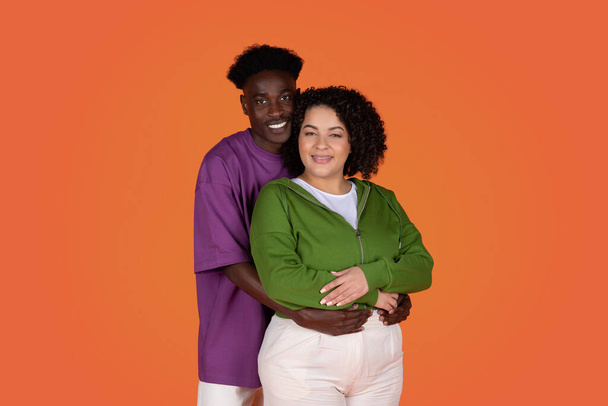 Studio portrait of cheerful beautiful young loving mixed race couple in casual hugging and smiling at camera on red background, african american guy embracing hispanic chubby woman, copy space - Foto, afbeelding