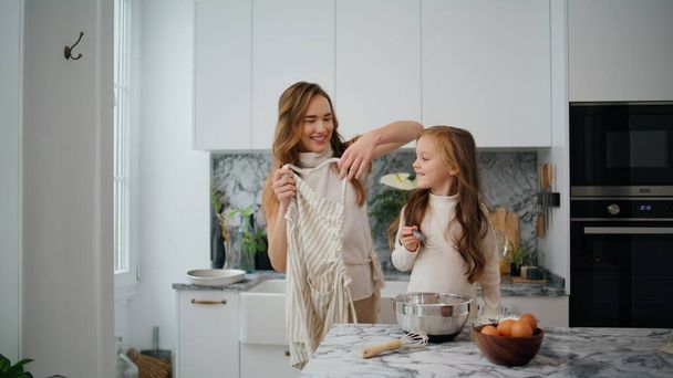 Cheerful woman putting apron on daughter light kitchen. Friendly family preparing cooking in apartment together. Single mother and baby girl spending time at home. Child having fun with female parent - Фото, изображение