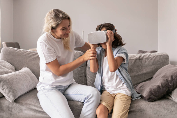 Μητέρα βοηθώντας το γιο του να φορούν ακουστικά εικονικής πραγματικότητας vr γυαλιά στο σαλόνι στο σπίτι έχοντας διασκέδαση που τέμνονται με εικονική πραγματικότητα παίζοντας παιχνίδια - Φωτογραφία, εικόνα