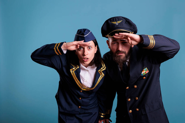 Πορτρέτο του καπετάνιου του αεροπλάνου και του ζεύγους αεροσυνοδών κοιτάζοντας την κάμερα, μέτρια λήψη. Ομάδα αεροσκαφών φορώντας στολή πτήσης, ενώ στέκεται στο στούντιο με μπλε φόντο - Φωτογραφία, εικόνα