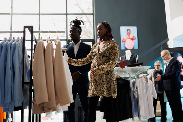 Έγκυος γυναίκα ψάχνει σε πουκάμισο υλικό ελέγχου σε εκθεσιακό χώρο, Αφροαμερικανός ζευγάρι ψώνια για ρούχα εγκυμοσύνης. Πελάτες που αγοράζουν εμπορεύματα μητρότητας σε κατάστημα ρούχων. Έννοια μόδας - Φωτογραφία, εικόνα
