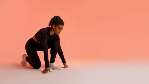 Motivated african american female runner, standing in crouch start position, ready for race over peach neon background, panorama. Fit black lady in fitwear preparing for running, free space - Foto, imagen