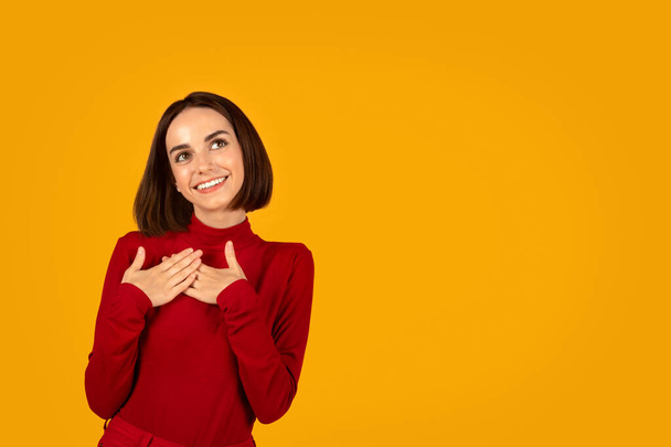 Emotional tranquil happy dreamy pretty young woman in red holding hands on chest, smiling and looking at copy space, lady dreaming about something or someone, orange studio background - Foto, afbeelding