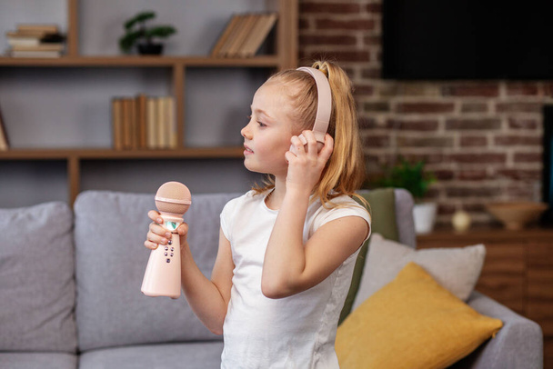 Happy little girl listening to music at home. Child wearing headphones using microphone, singing song on sofa in room. - Photo, Image