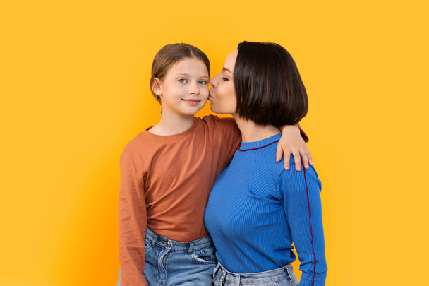 Loving Family. Young Mother Kissing Her Happy Little Daughter, Smiling Mom And Preteen Female Child Embracing And Having Fun Together, Bonding Over Yellow Studio Background, Copy Space - Foto, afbeelding