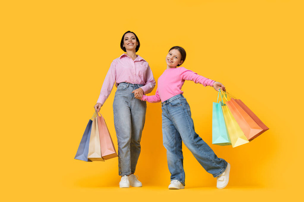 Family Shopping. Happy Young Mother And Little Daughter Posing With Lots Of Bright Shopper Bags, Cheerful Mom And Preteen Female Child Holding Hands And Standing Over Yellow Background, Copy Space - Foto, Imagen