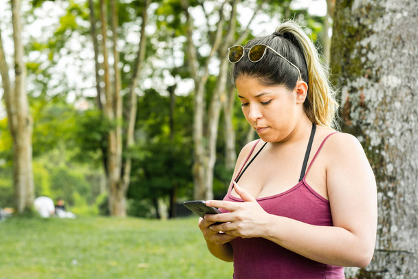 beautiful young latina woman, next to a big tree in a park looking at her cell phone - Photo, image