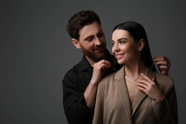 Man putting elegant necklace on beautiful woman against dark grey background - Fotó, kép