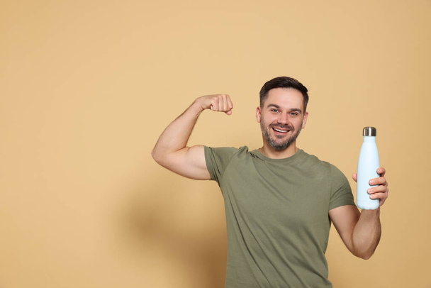 Happy man holding thermo bottle and showing arm on beige background, space for text - Fotoğraf, Görsel