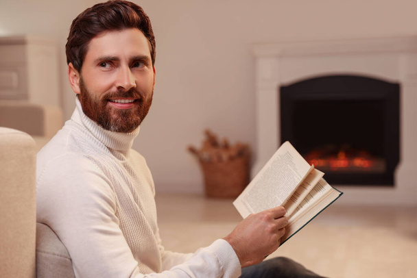 Handsome man reading book near fireplace in room - Foto, imagen