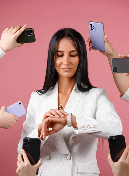 A HAPPY BUSINESS WOMAN IN A WHITE JACKET AND BLACK HAIR IS LOOKING AT THE CLOCK SURROUNDED BY MANY MODELS OF PHONES ON PINK BACKGROUND. High quality photo - Foto, imagen