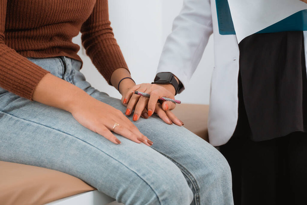 a female doctor in doctors coat put her hand above the hand of the female patient in brown clothes and denim pants - Foto, immagini