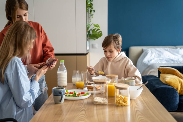 Μητέρα και κόρη χρησιμοποιούν το κινητό τηλέφωνο στο τραπέζι της κουζίνας ενώ παίρνουν πρωινό μαζί με την οικογένεια. Έφηβη που δείχνει κάτι στη μαμά στο κινητό την ώρα του φαγητού. Χρήση κινητού τηλεφώνου και οικογενειακός δεσμός - Φωτογραφία, εικόνα