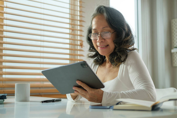 Щаслива старша жінка в окулярах насолоджується переглядом інтернету на цифровому планшеті в затишному будинку
. - Фото, зображення