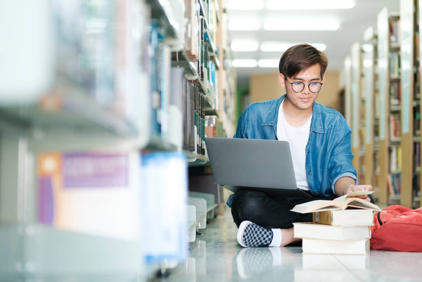 Young male college, highschool student wearing eyeglasses and casual clothings sitting on the floor reading book, studying and doing research using laptop for school project at a library. E-Learning and Educational concept. - Foto, imagen