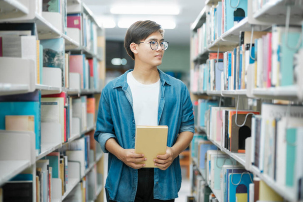 Νεαρός φοιτητής με casual ντύσιμο με σακίδιο πλάτης που στέκεται, διαβάζει ή επιλέγει βιβλία για να διαβάσει στη βιβλιοθήκη για σπουδές, ακαδημαϊκή έρευνα ή σχολικές εργασίες. Μάθηση, Εκπαίδευση έννοια. - Φωτογραφία, εικόνα