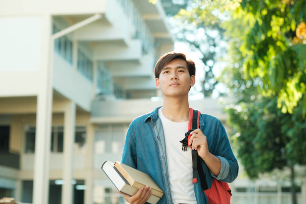 Young smart motivated male collage student in casual clothings and backpack standing outdoor with campus building in the backfround and holding books while looking up ready for study. Education concept. - 写真・画像