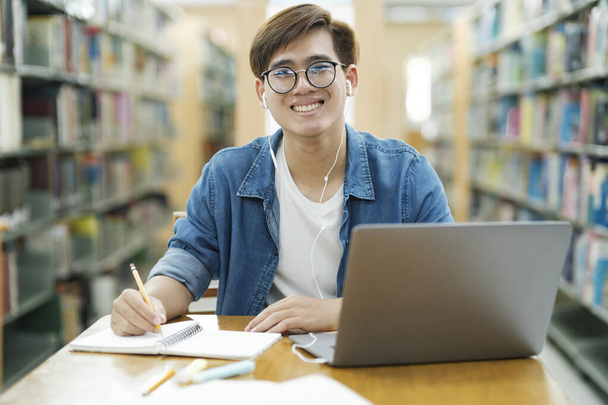 Młody student college 'u w okularach i w luźnych ciuchach, siedzący przy biurku, studiujący, czytający książki i zapisujący notatki przy użyciu laptopa, noszący słuchawki patrzące na kamerę w bibliotece. - Zdjęcie, obraz