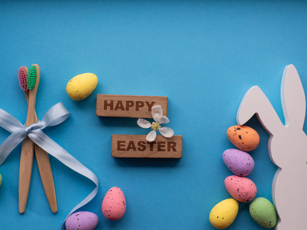 Happy Easter and dentist concept. Wooden toothbrushes with Easter decorations on blue background. Dentist Easter greating card. Top view, flat lay. Bamboo toothbrushes, colorful eggs, white bunny. - Photo, Image
