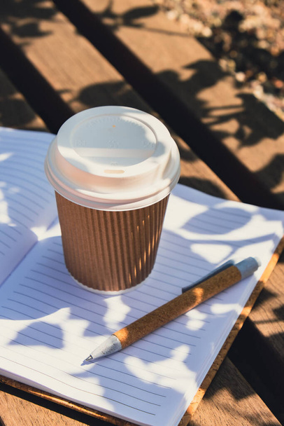 Eco recycling paper cup with coffee or tea on kraft paper with empty paper notebook on wooden bench. Concept of study work outdoors. Take away coffee to go. Copy space for text. Disposable Cardboard - Photo, Image