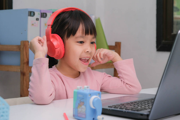 Χαριτωμένο κορίτσι δημοτικού σχολείου φορώντας ακουστικά και χρησιμοποιώντας έναν φορητό υπολογιστή. Ευτυχισμένα παιδιά από την Ασία σπουδάζουν online διαδραστικά με φορητό υπολογιστή ή κατ 'οίκον διδασκαλία, ακούγοντας μουσική ή παίζοντας παιχνίδια. - Φωτογραφία, εικόνα