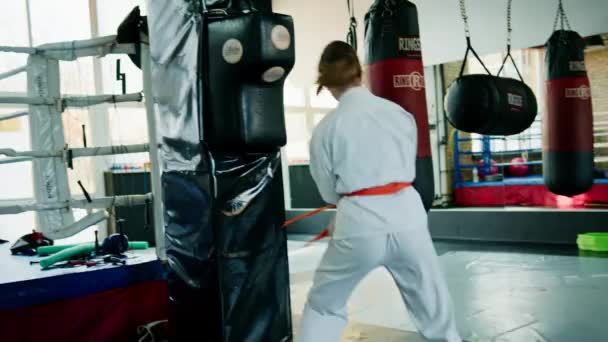Ένα κορίτσι με κιμονό γυμνάζεται με σάκο του μποξ στο γυμναστήριο ενώ μαθαίνει καράτε στις πολεμικές τέχνες. - Πλάνα, βίντεο