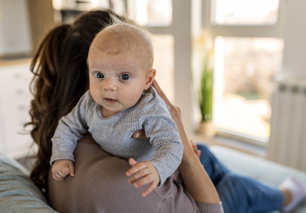 Portret uroczego niemowlęcia na ramieniu matki patrzącego w kamerę. Matczyna koncepcja miłości i opieki - Zdjęcie, obraz