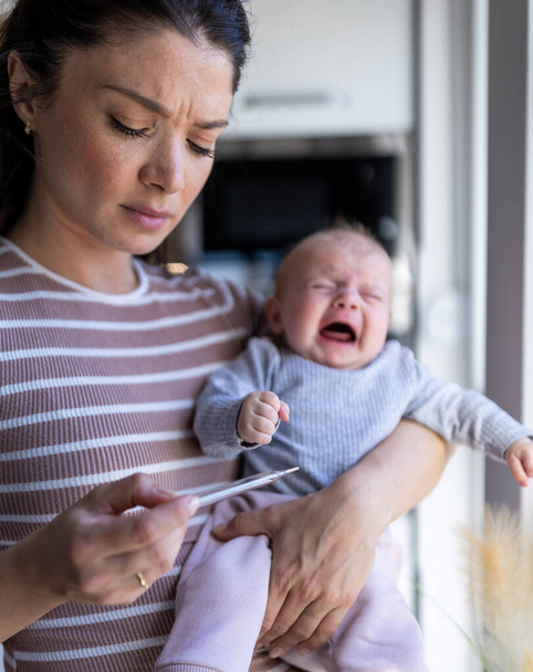 Zaniepokojona matka trzymająca płaczące dziecko w jednej ręce w domu i patrząca na termometr z powodu gorączki - Zdjęcie, obraz