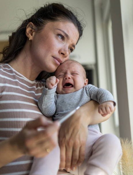 Huolestunut äiti, jolla on itkevä vastasyntynyt vauva ja lämpömittari toisessa kädessä kotona. Äitiys rakkaus ja hoito käsite - Valokuva, kuva