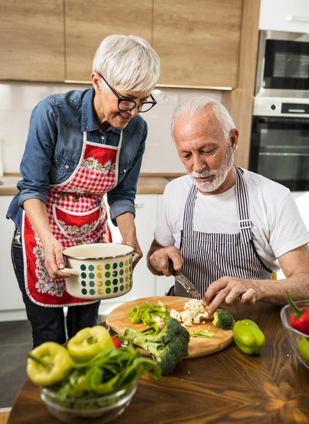 Starsza para przygotowuje zupę, mąż kroi warzywa, a żona trzyma garnek - Zdjęcie, obraz