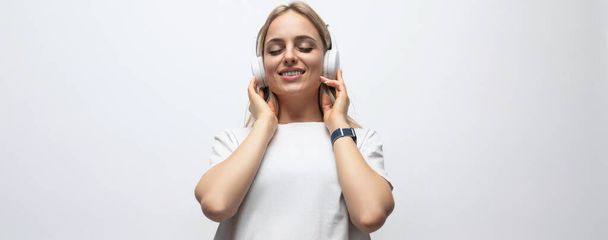 Beyaz atletli genç bir kadının beyaz arka planda kablosuz kulaklıkla podcast dinlerken çekilmiş yatay fotoğrafı.. - Fotoğraf, Görsel