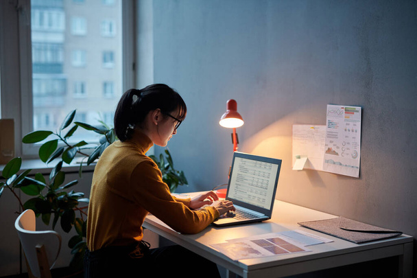 Giovane donna d'affari seduta sul posto di lavoro e che lavora con il progetto online utilizzando il computer portatile, lavora fino a tardi in ufficio - Foto, immagini