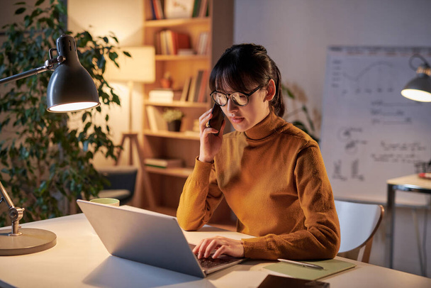 Молода бізнес-леді в окулярах розмовляє на мобільному телефоні під час роботи за столом з ноутбуком під час перебування в офісі
 - Фото, зображення