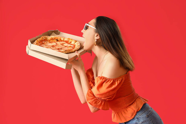 Beautiful woman in sunglasses eating tasty pizza on red background - 写真・画像