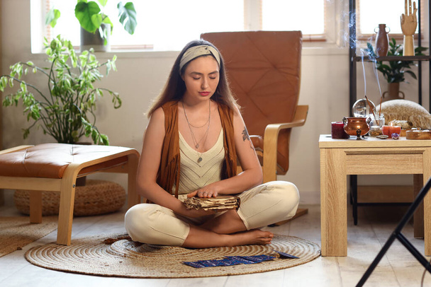 Tarot kartları ve büyü kitabı olan genç cadı evde meditasyon yapıyor. - Fotoğraf, Görsel