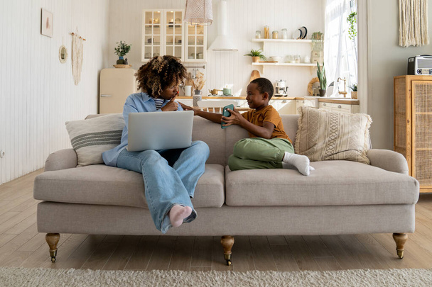 Äitiys- ja etätyö. Kiireinen afroamerikkalainen äiti freelancer istuu sohvalla kannettavan tietokoneen kanssa pyytäen pientä poikaa olemaan hiljaa kokouspuhelun aikana, yrittäen hallita etätyötä ja lastenhoitoa. - Valokuva, kuva